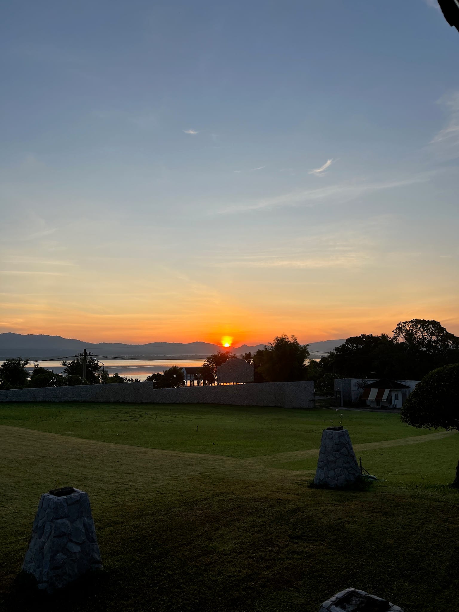 Sunset view from a lawn with low stone lantern pillars, looking over a white perimeter wall toward a large body of water with hills on the horizon.