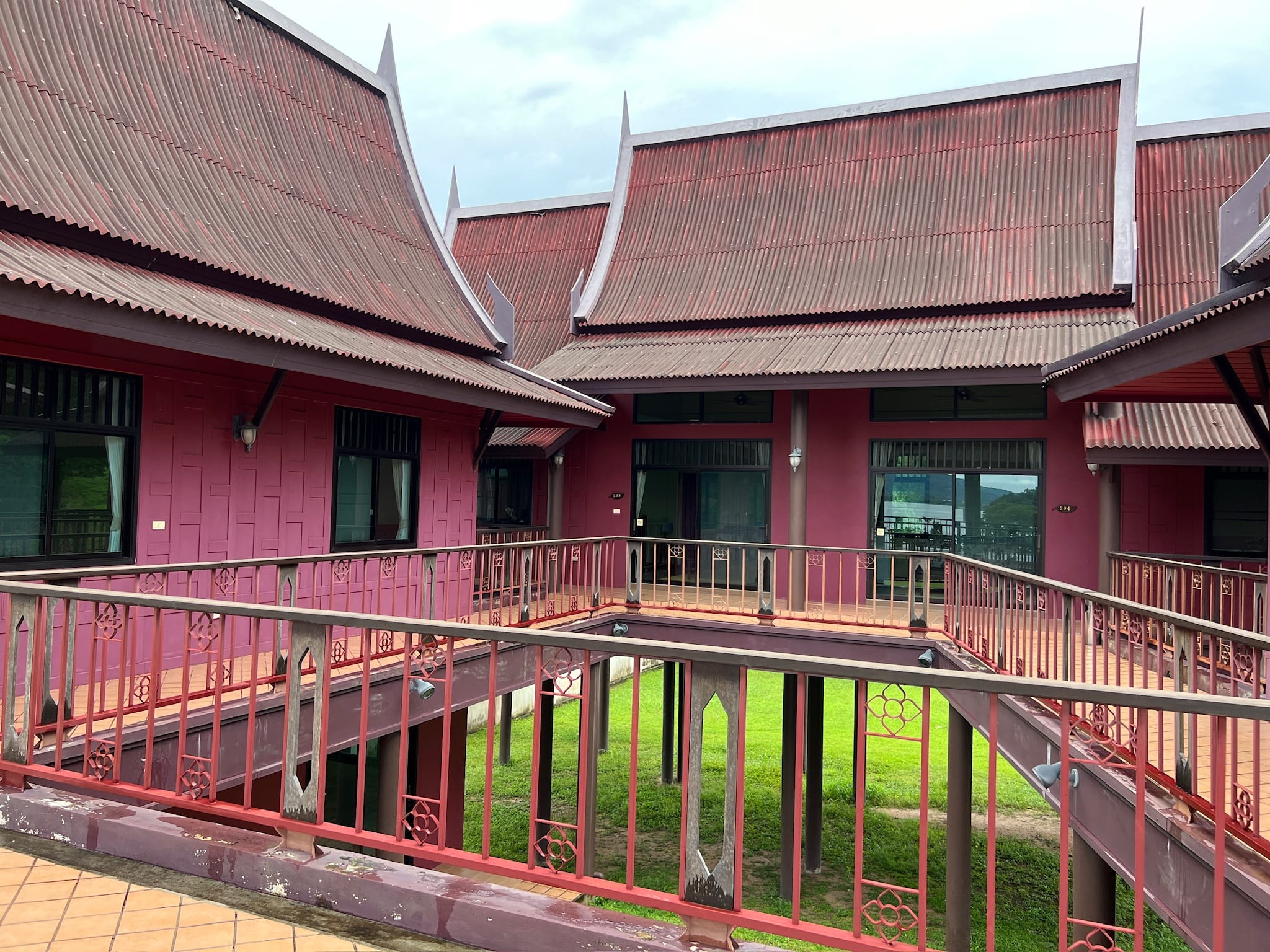 Upper-level walkway of a dark red traditional Thai-style building, with ornamental red metal railings surrounding an open lightwell over a grass courtyard below.