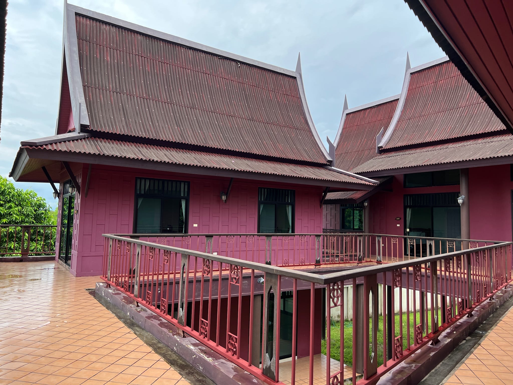 Exterior terrace of a dark red Thai-style house showing two steep pointed gable roofs with corrugated sheeting, dark-framed windows, and a terracotta-tiled deck.
