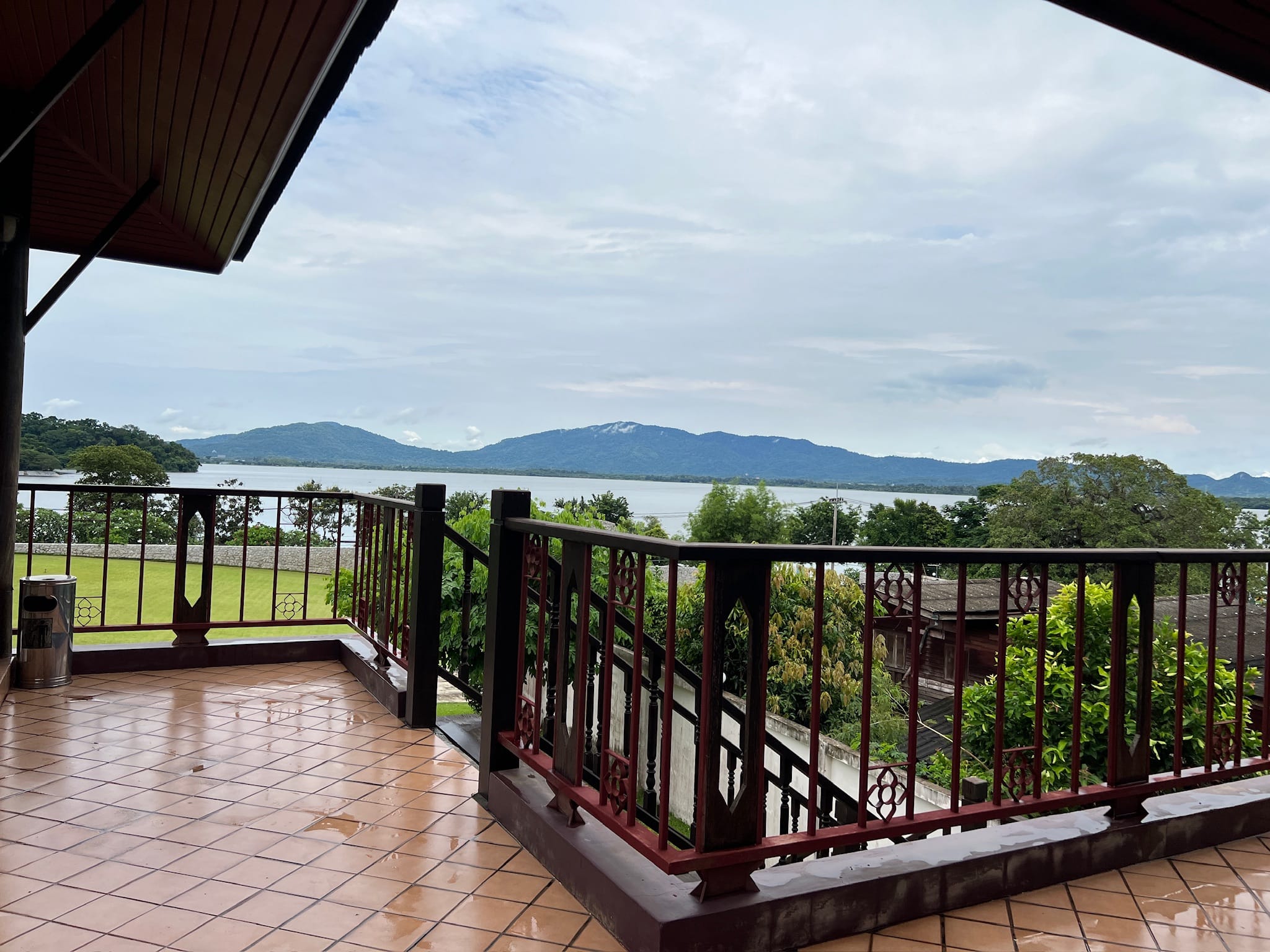 View from a covered tiled balcony with dark wood and red ornamental railings, overlooking a lawn, treetops, and a wide lake ringed by green hills.