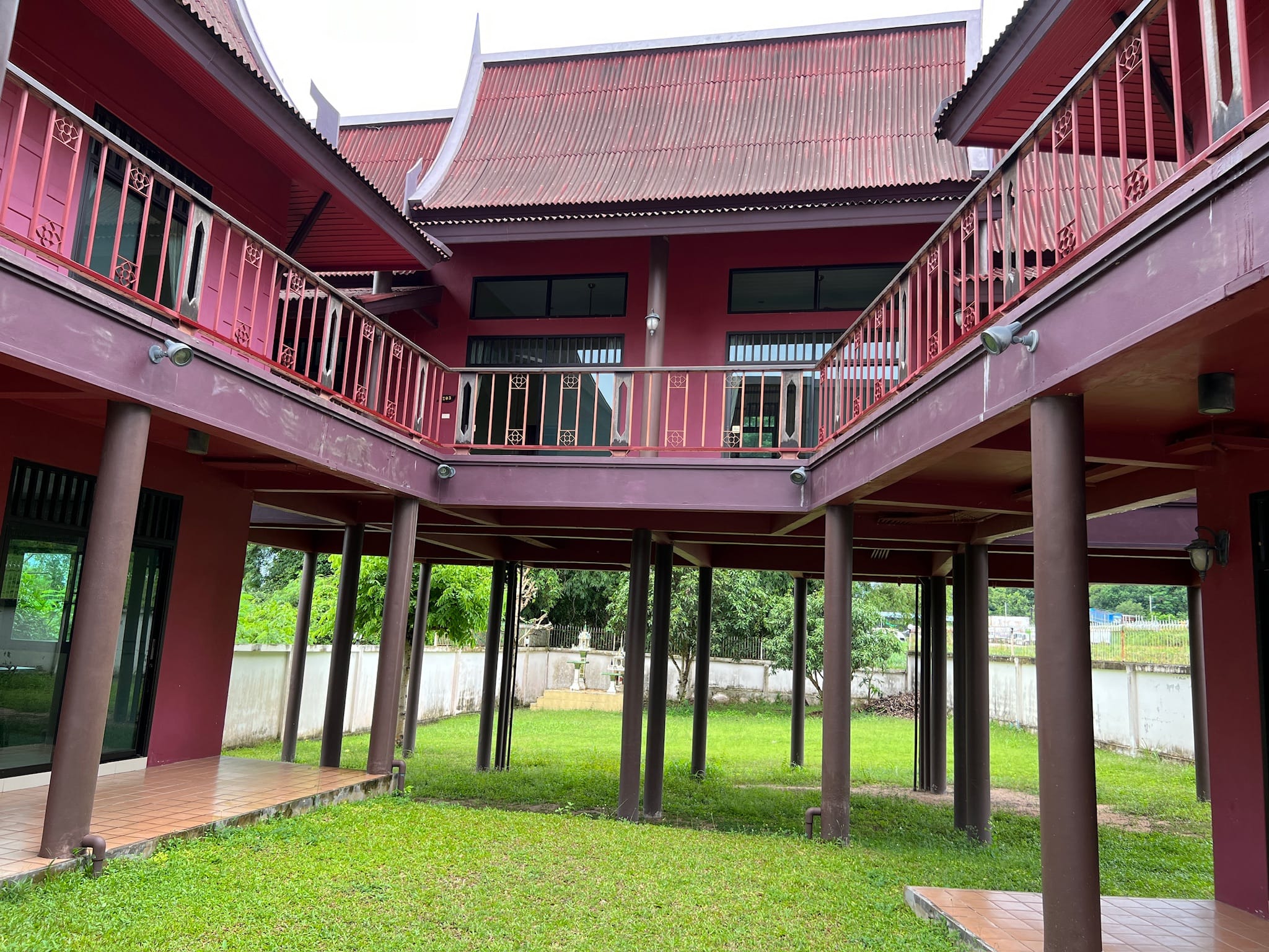 Grassy inner courtyard of a two-storey dark-red traditional Thai-style house raised on round stilts, with an L-shaped upper balcony and ornamental railings.