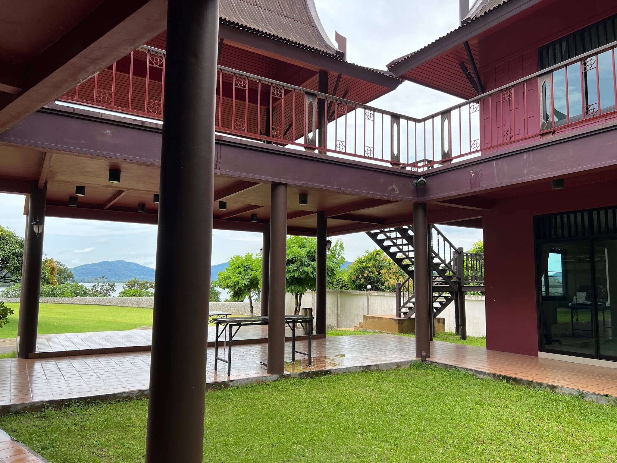 Shaded ground-level area beneath a traditional Thai-style house on dark stilts, with a tiled patio, folding tables, and a wooden staircase to the upper balcony.