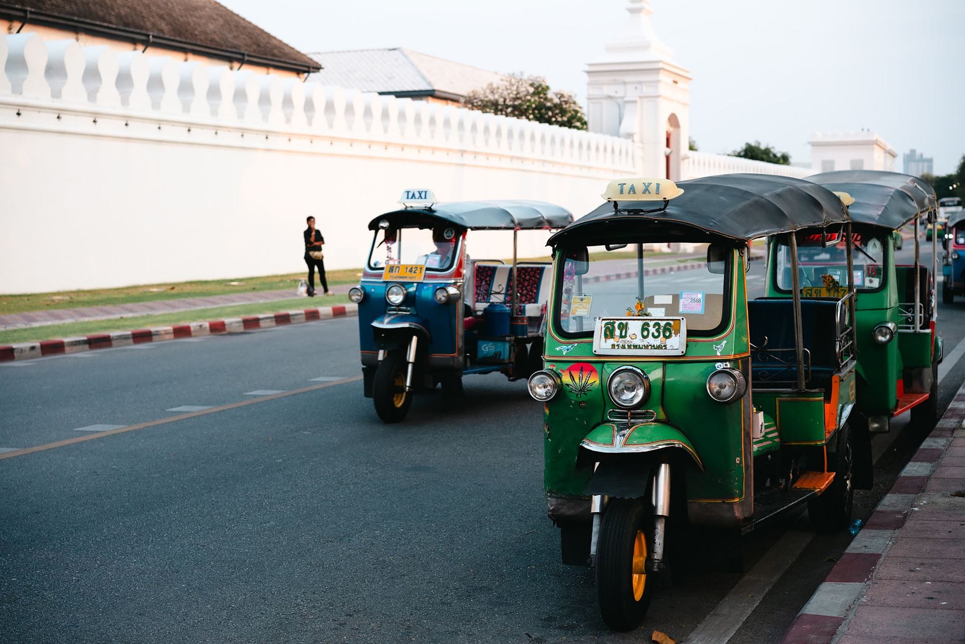 Green and blue tuk tuk taxis lined up along the white wall of the Grand Palace in Bangkok